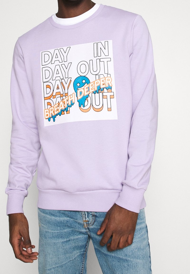 Lila sweatshirt med en vit grafik som har orange och blå text. Designen inkluderar orden "DAY IN DAY OUT" och en blå karaktär.