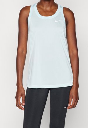 Femme portant un haut sans manches Nike bleu clair et des leggings noirs Nike, debout devant un fond uni.