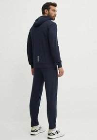 Tuta blu navy con cappuccio, caratterizzato da una vestibilità aderente e dettagli riflettenti sulla schiena. Realizzato in tessuto morbido e con texture per il massimo comfort.