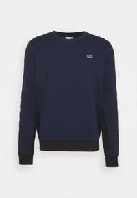 Sweat-shirt bleu marine à manches longues avec encolure ronde, panneaux latéraux noirs, poignets et ourlet côtelés, et petit logo crocodile vert sur la poitrine.