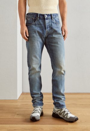 Svetle modré džíny z denimu s ošoupaným vzhledem, rovný střih a design s pěti kapsami, v kombinaci se šedými a bílými sportovními botami.