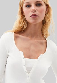 Valkoinen pitkähihainen paita, jossa on avara kaula-aukko ja etupaneelin napitus, käytetty valkoisen ribbitekstuurin tanktopin päällä, pehmeä rakenne.