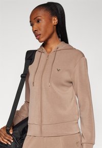 Felpa con zip beige realizzata in tessuto morbido, con cappuccio, tasche frontali e una spilla a forma triangolare con accessori dorati.