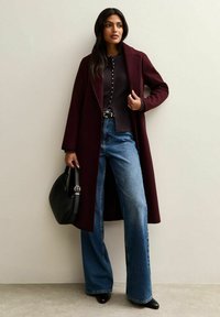 Cappotto lungo bordeaux sopra un maglione scuro, con jeans blu a gamba larga. Completato da scarpe nere a punta e una borsa nera.