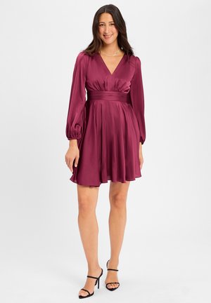 Burgundy satin dress with a V-neck, long puff sleeves, cinched waist, and flared skirt. Black strappy heels complete the look.