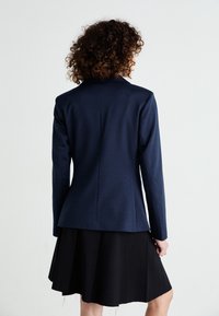 Femme aux cheveux bouclés portant un blazer bleu marine et une jupe plissée noire, debout face à un fond gris clair uni.