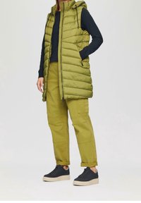 Gilet imbottito verde oliva con cappuccio, texture trapuntata, chiusura con zip e tasche laterali, indossato su una maglia scura a maniche lunghe e pantaloni cargo.