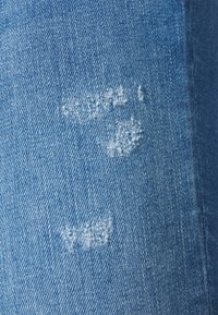 Tejido de mezclilla azul con pequeñas manchas blancas desgastadas y costuras visibles en el borde derecho.