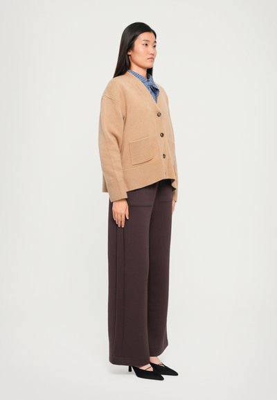 Beige kötött kardigán négy gombbal és két elülső zsebbel, sötétbarna, bő szárú nadrággal és fekete, hegyes orrú magassarkú cipővel párosítva.