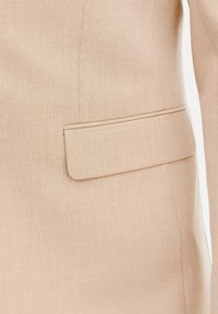 Beżowa marynarka o dopasowanym kroju, wykonana z gładkiego materiału; posiada kieszeń w talii oraz staranne przeszycia wzdłuż krawędzi. Prosty i zorganizowany design.