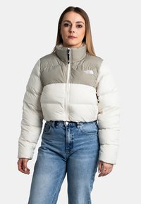 Giacca corta e voluminoso in beige chiaro e crema con colletto alto, chiusura con zip e dettaglio del logo sul petto; abbinata a jeans blu.