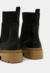Bottines en cuir noir avec un dessus en textile noir côtelé, présentant une semelle épaisse beige et un accent logo doré à l'arrière.