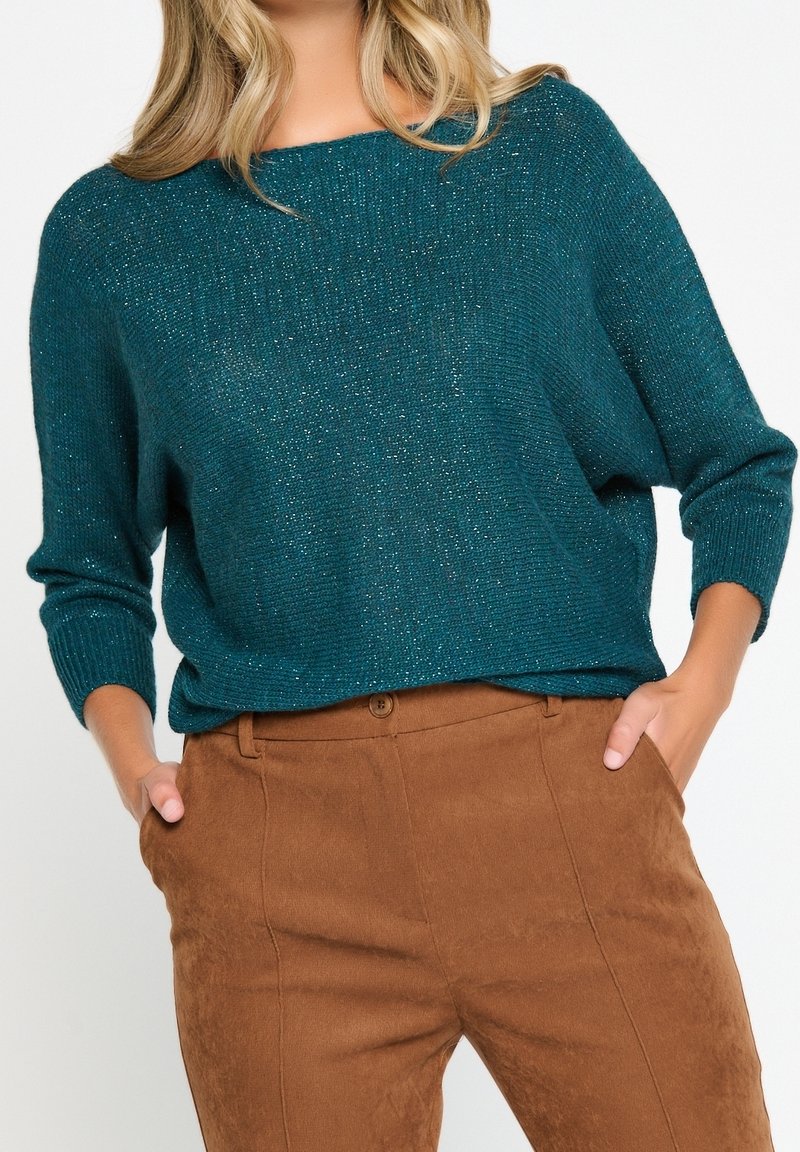 Teal stickad tröja med metalltrådar, rund halsringning och trekvartslånga ärmar, parat med bruna mockabyxor.