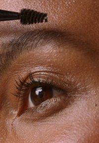 Un cepillo de cejas con una cabeza de cerdas en espiral está sostenido cerca de un ojo marrón, revelando largas pestañas y una piel suave y texturada.
