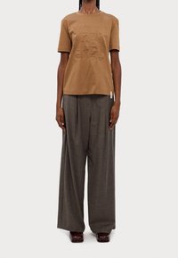 T-shirt in cotone marrone con design embossato e maniche corte, abbinato a pantaloni a gamba larga grigi realizzati in tessuto texturizzato, indossato con scarpe scure.