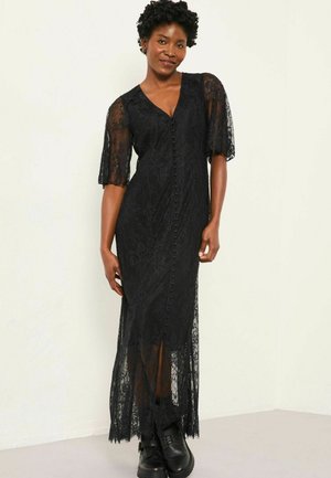 Schwarzes Spitzenkleid mit kurzen, schwingenden Ärmeln, knöpfbarem Vorderteil und langem Saum, das eine durchsichtige Textur und seitliche Schlitze aufweist. Getragen mit schwarzen Stiefeln.