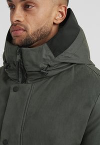 Dunkelgrüne Jacke mit hohem Kragen, Frontreißverschluss und Knopfverschluss. Verfügt über Zugbandversteller und eine glatte, matte Textur.