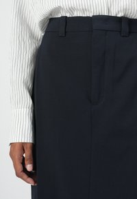Pantalon bleu marine foncé avec une taille plate, passants de ceinture et poches latérales, associé à une chemise blanche rayée à manches longues. Détail en tissu texturé visible.