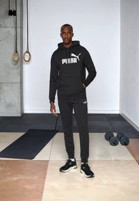 Schwarzer Kapuzenpullover und passende Jogginghose mit weißem Puma-Logo. Das Modell trägt schwarze Sportschuhe. Der Hintergrund zeigt Fitnessgeräte.