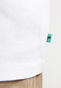 T-shirt en coton blanc avec un ourlet latéral orné d'une étiquette turquoise. Texture lisse et design simple. Couture visible à l'ourlet inférieur.
