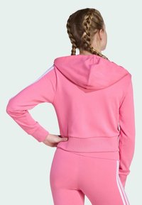 Sudadera rosa hecha de tela suave con capucha, que presenta franjas blancas en las mangas y un diseño ajustado.