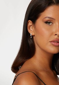 Silberne Creolen mit einem kleinen Tropfenanhänger, die ein strukturiertes Design und funkelnde Akzente aufweisen, getragen vor einem glatten Hautton.