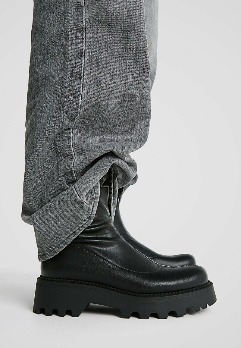 Bottes en cuir noires épaisses portées avec un jean gris ample sur un fond clair uni.