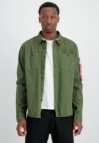 Camisa verde de botones hecha de tela, con dos bolsillos en el pecho, un bolsillo con cremallera en la manga y una etiqueta de tela roja con texto.