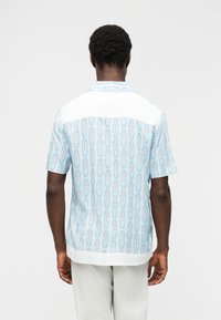 Άνδρας που φοράει ανοιχτό μπλε πουκάμισο με κοντά μανίκια και λευκό γιακά και τελείωμα, συνδυασμένο με ανοιχτό γκρι παντελόνι, στέκεται μπροστά σε απλό λευκό φόντο.