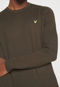 Pull en tricot vert olive avec un col rond et des poignets côtelés, arborant un petit logo brodé jaune sur le côté gauche de la poitrine.