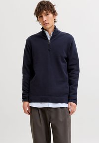 Μπλε ναυτικό πουλόβερ με γιακά μισού φερμουάρ, υφή υφάσματος, άνετη γραμμή και ριμπ μανσέτες. Φορεμένο πάνω από ένα ανοιχτό μπλε ριγέ πουκάμισο και γκρι παντελόνι.