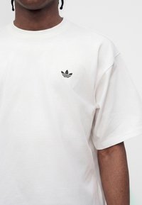 Άτομο που φοράει ένα απλό λευκό T-shirt Adidas με μικρό μαύρο λογότυπο Adidas trefoil στο αριστερό στήθος.