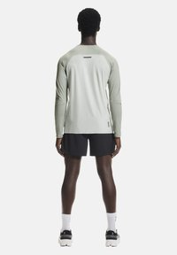 Maglietta sportiva a maniche lunghe di colore verde chiaro con tessuto strutturato, abbinata a pantaloncini neri. Presenta un logo discreto sul retro e dettagli sui polsini.