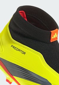 Botín de fútbol amarillo brillante con acentos en negro y rojo, que presenta un alto cuello tipo calcetín, superficie texturizada y un atrevido logo "PREDATOR".