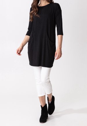 Túnica negra con mangas tres cuartos, dos grandes bolsillos laterales, combinada con pantalones cortos blancos y botines negros. Tejido suave, corte relajado.