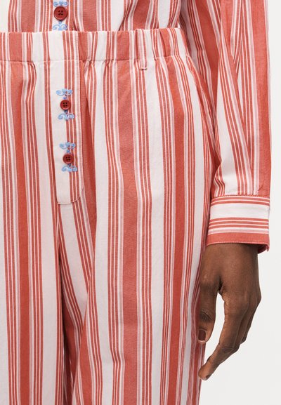 Κοντινό πλάνο ενός ατόμου που φοράει κόκκινο και άσπρο παντελόνι με κάθετες ρίγες και αντίστοιχο μακρυμάνικο πουκάμισο με κόκκινα κουμπιά και ανοιχτομπλε κέντημα.