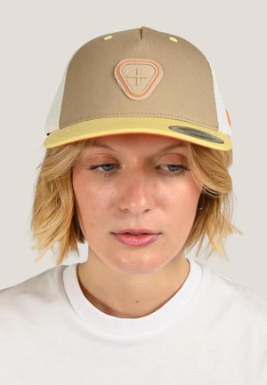 Femme blonde portant une casquette de baseball beige et jaune avec un logo en forme de signe plus, regardant vers le bas, vêtue d'une chemise blanche.