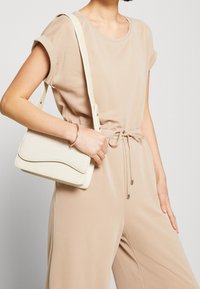 Witte geweven handtas met een gebogen bovenkant, synthetisch materiaal, schouderriem, vastgehouden door een persoon die een beige outfit met een trekkoord taille draagt.