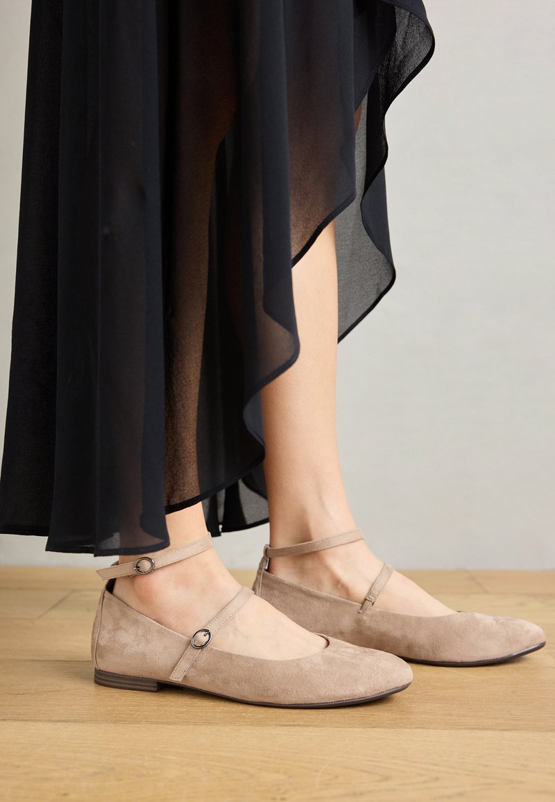 Beige Wildlederballerinas mit mandelförmiger Spitze und Knöchelriemen mit einem runden Akzent, getragen zu einem durchsichtigen schwarzen Rock, auf einem Holzfußboden.