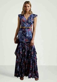 Mujer luciendo un vestido maxi sin mangas azul con estampado floral y volantes, un cinturón beige, sosteniendo un pequeño clutch y usando sandalias.