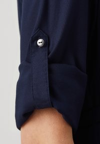 Námořnická modrá košile má manžetu s stříbrným tlačítkem a dírkou na knoflík, hladkou texturu a design ohrnutého rukávu pro pohodlný fit.