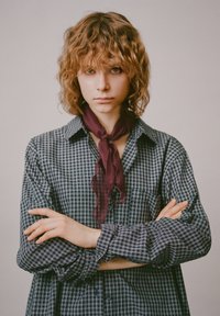 Koszula z długim rękawem w granatowo-białą kratę, z kołnierzykiem na guziki, w zestawieniu z głębokim bordowym, marszczonym szalikiem na szyję. Modelka ma falowane włosy.