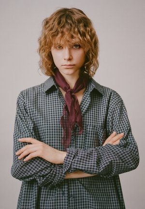 Košeľa s dlhým rukávom, námornícka modrá a biela károvaná so zapínaním na gombíky na golieri, doplnená hlboko burgundským šálom s volánikmi okolo krku. Modelka má vlnité vlasy.