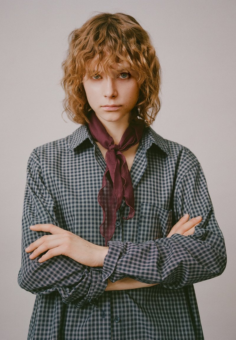 Koszula z długim rękawem w granatowo-białą kratę, z kołnierzykiem na guziki, w zestawieniu z głębokim bordowym, marszczonym szalikiem na szyję. Modelka ma falowane włosy.