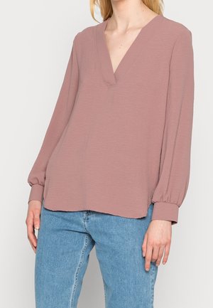 Femme portant un chemisier mauve à manches longues avec col en V et manches retroussées, associé à un jean bleu clair, sur un fond uni.