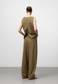 Kvinna med brunt hår i en knut, klädd i en ärmlös olivgrön topp med slits i ryggen och vida matchande byxor, stående mot en enhetlig bakgrund.