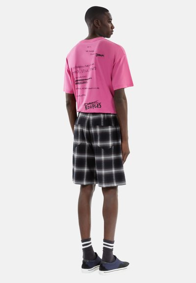 Maglietta grafica rosa con stampa nera, abbinata a pantaloni corti a quadri bianchi e neri. Il modello indossa scarpe nere e calzini a strisce.