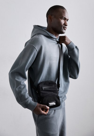Sacoche noire en bandoulière avec des accents en maille, plusieurs poches zippées et un logo. Porte avec un sweat à capuche gris et un pantalon assorti.