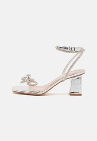Sandalia de tacón alto plateada con punta cuadrada, material transparente, correa adornada y un tacón de bloque grueso y espejo. Incluye un cristal decorativo.