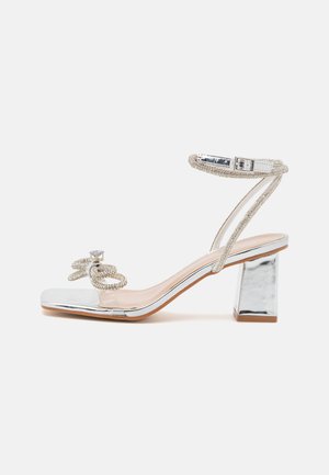 Zilveren hoge hak sandaal met een vierkante neus, transparant materiaal, versierde band en een chunky, spiegelende blokhak. Bevat een decoratieve kristal.